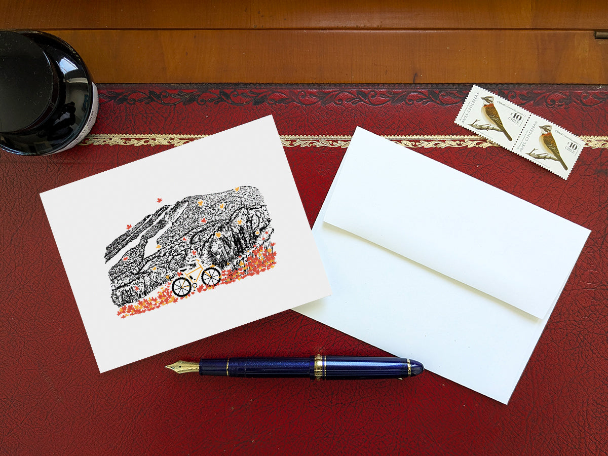 Greeting card with image of a mountain bike surrounded by brightly colored leaves with a mountain in the background on a table with blue pen and envelope and stamps.