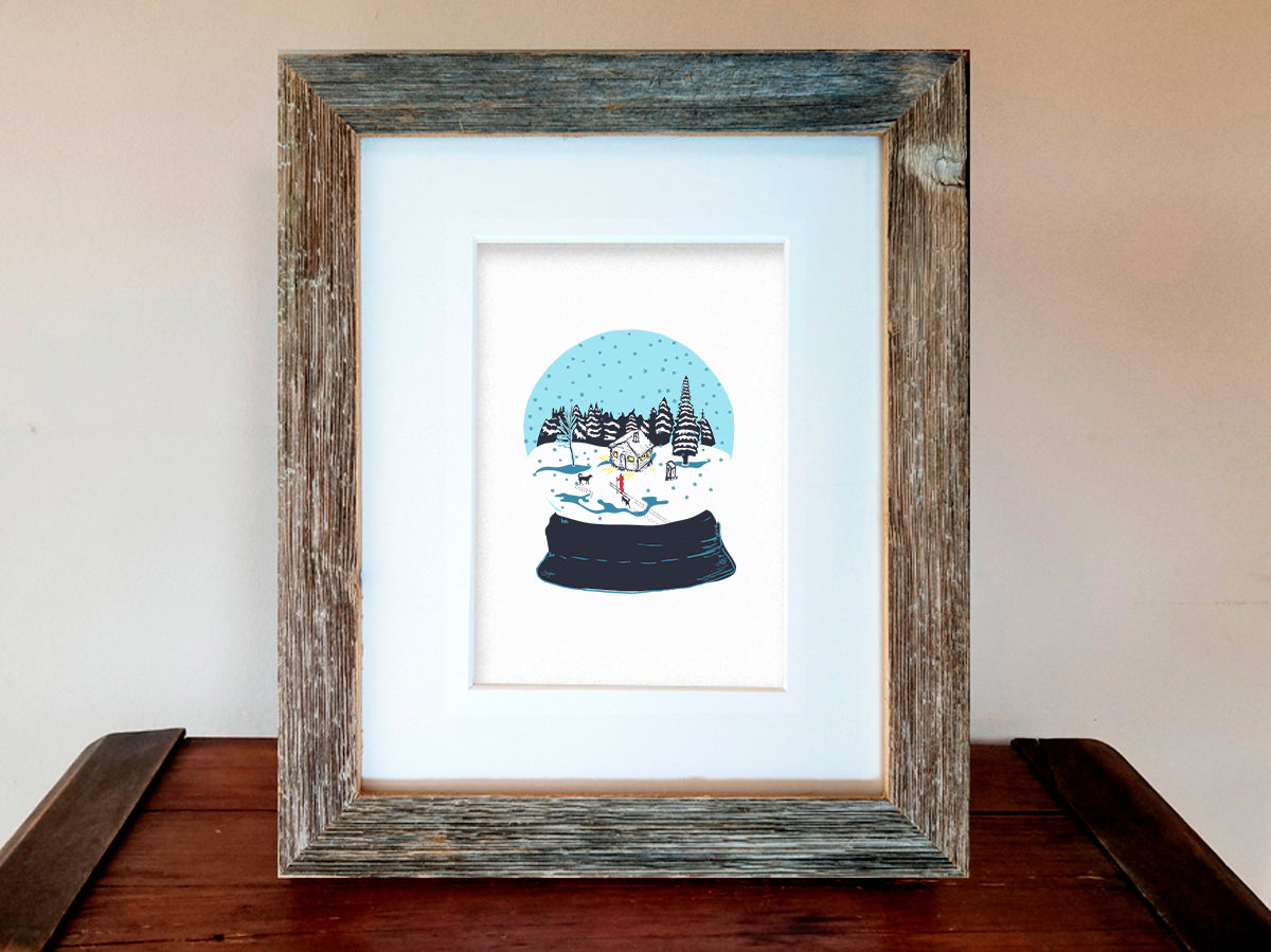 A framed art print featuring a snow globe illustration with a cabin, skier, and two dogs among a snowy landscape, displayed on a wooden shelf.