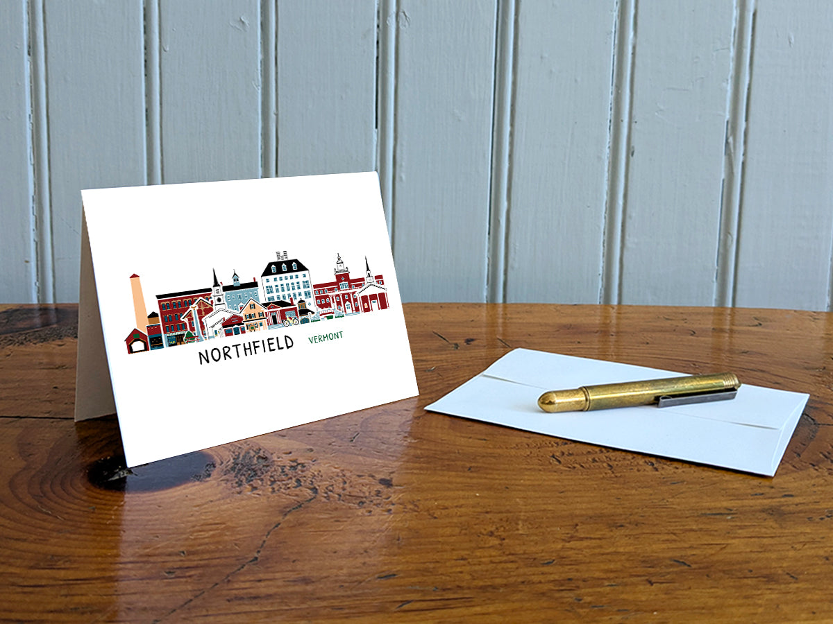 Greeting card with image of buildings arranged in a city skyline with the text Northfield Vermont on a table with gold pen and envelope.