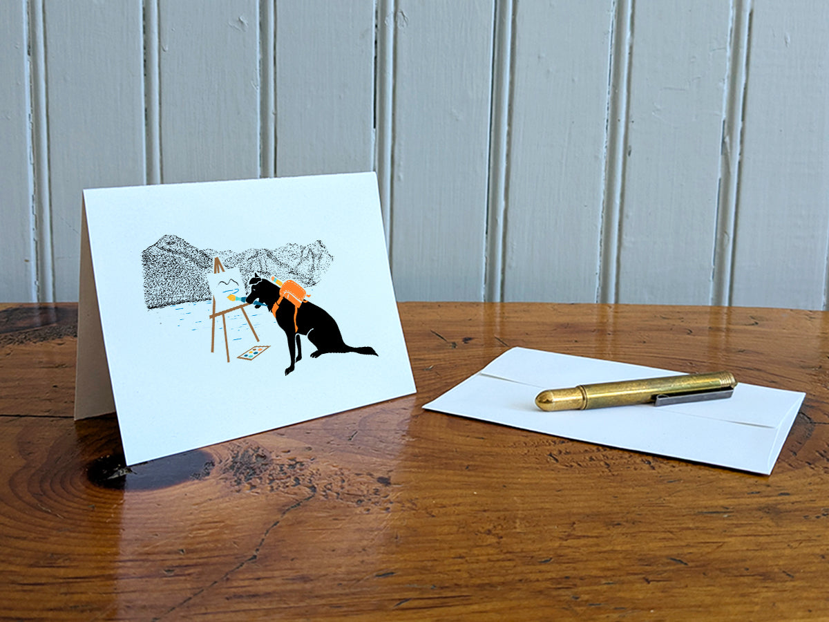 White card with drawing of dog painting in the mountains on a wooden surface with a pen and envelope.