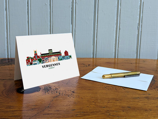 Greeting card with image of buildings arranged in a city skyline with the text Vergennes Vermont on a table with gold pen and envelope.