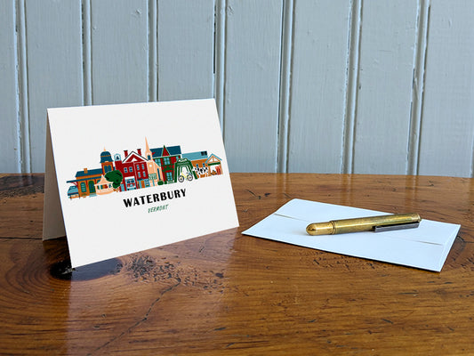 Greeting card with image of buildings arranged in a city skyline with the text Waterbury Vermont on a table with gold pen and envelope.