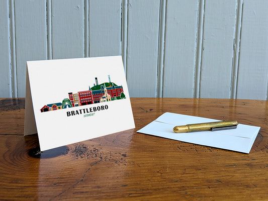 Greeting card with image of buildings arranged in a city skyline with the text Brattleboro Vermont on a table with gold pen and envelope.