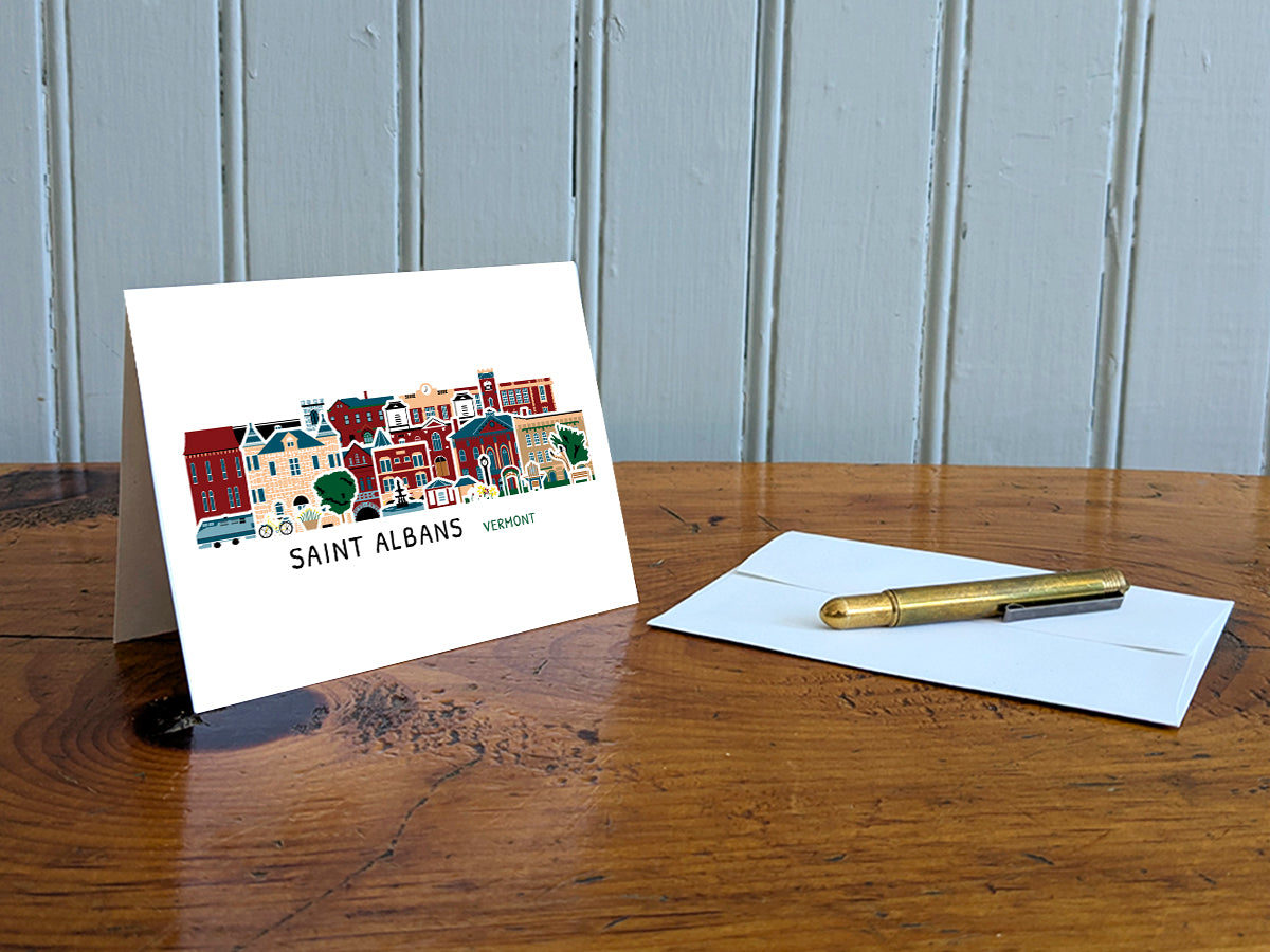 Greeting card with image of buildings arranged in a city skyline with the text Saint Albans Vermont on a table with gold pen and envelope.