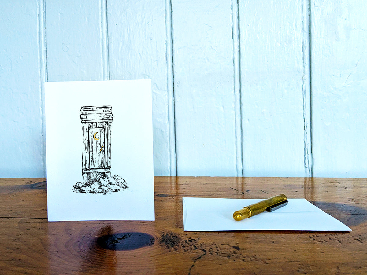 White card with a black ink drawing of an outhouse on a wooden surface with a pen and envelope.