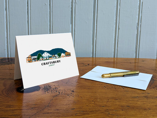 Greeting card with image of buildings arranged in a city skyline with the text Craftsbury Vermont on a table with gold pen and envelope.