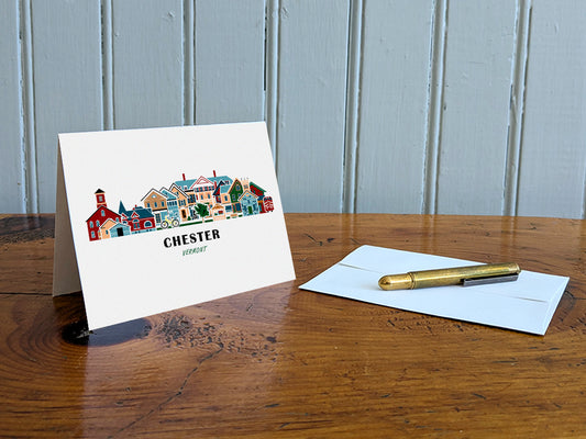 Greeting card with image of buildings arranged in a city skyline with the text Chester Vermont on a table with gold pen and envelope.