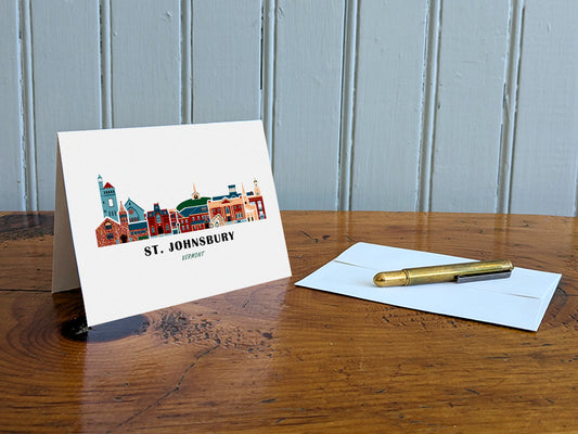 Greeting card with image of buildings arranged in a city skyline with the text St. Johnsbury Vermont on a table with gold pen and envelope.