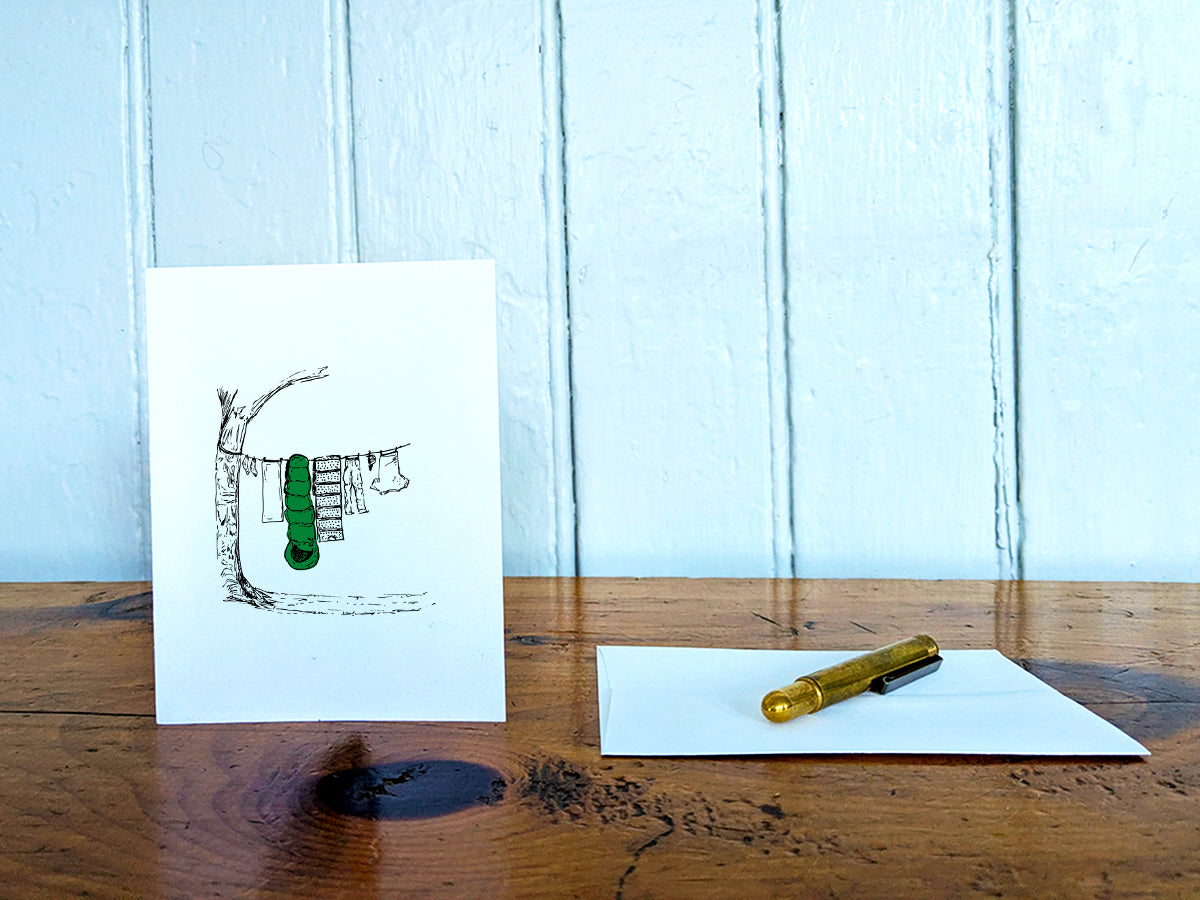 White card with a black ink drawing of laundry hanging from a tree on a wooden surface with a pen and envelope.