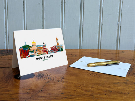 Greeting card with image of buildings arranged in a city skyline with the text Montpelier Vermont on a table with gold pen and envelope.