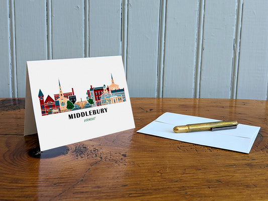 Greeting card with image of buildings arranged in a city skyline with the text Middlebury Vermont on a table with gold pen and envelope.