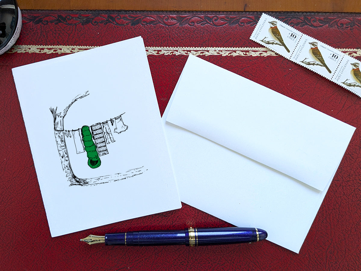 White card with a black ink drawing of laundry hanging from a tree on a red surface with a pen, envelope and stamp.
