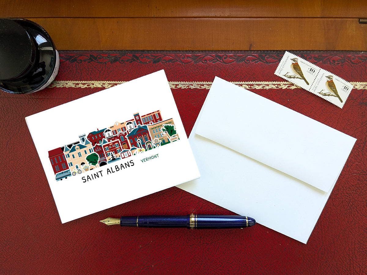 Greeting card with image of buildings arranged in a city skyline with the text Saint Albans Vermont on a red table with blue pen, stamps and envelope.