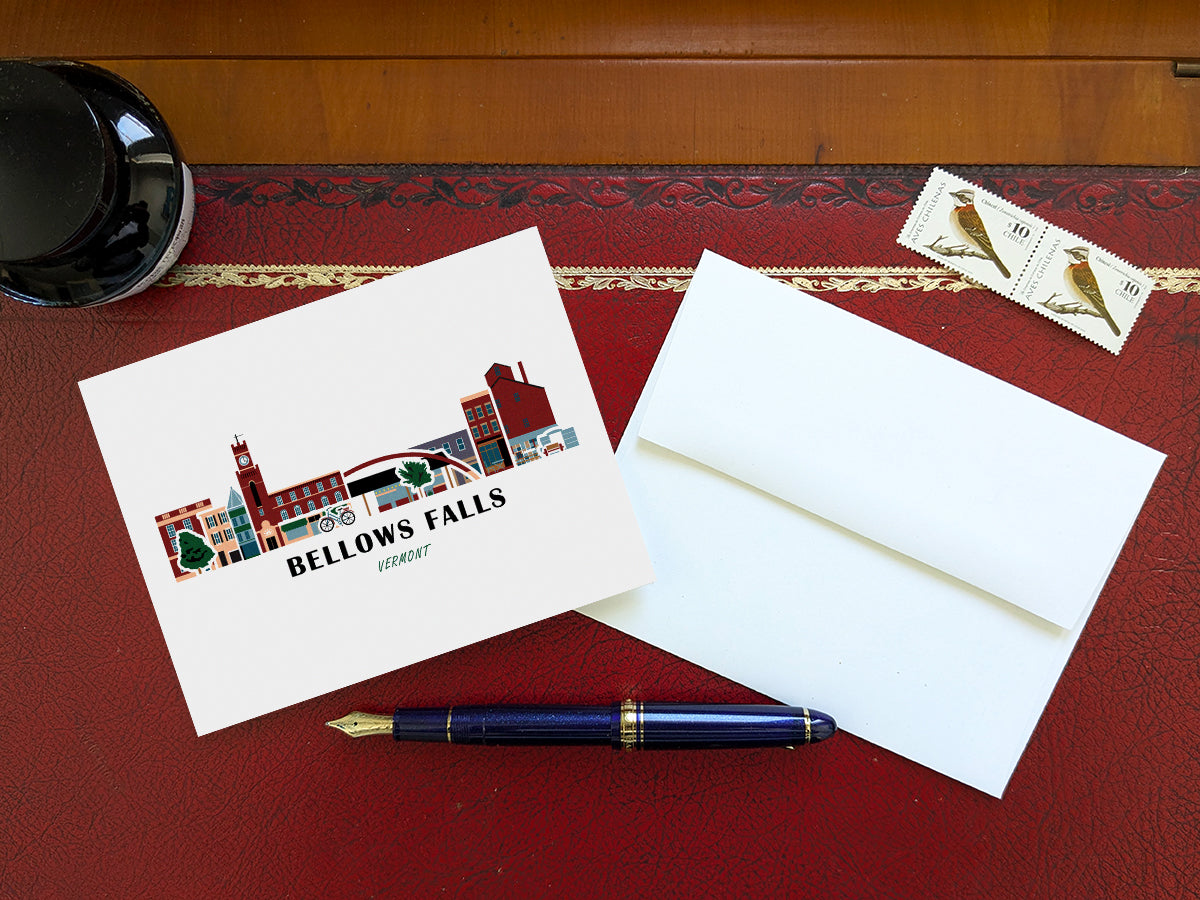 Greeting card with image of buildings arranged in a city skyline with the text Bellows Falls Vermont on a table with blue pen and envelope and stamps.