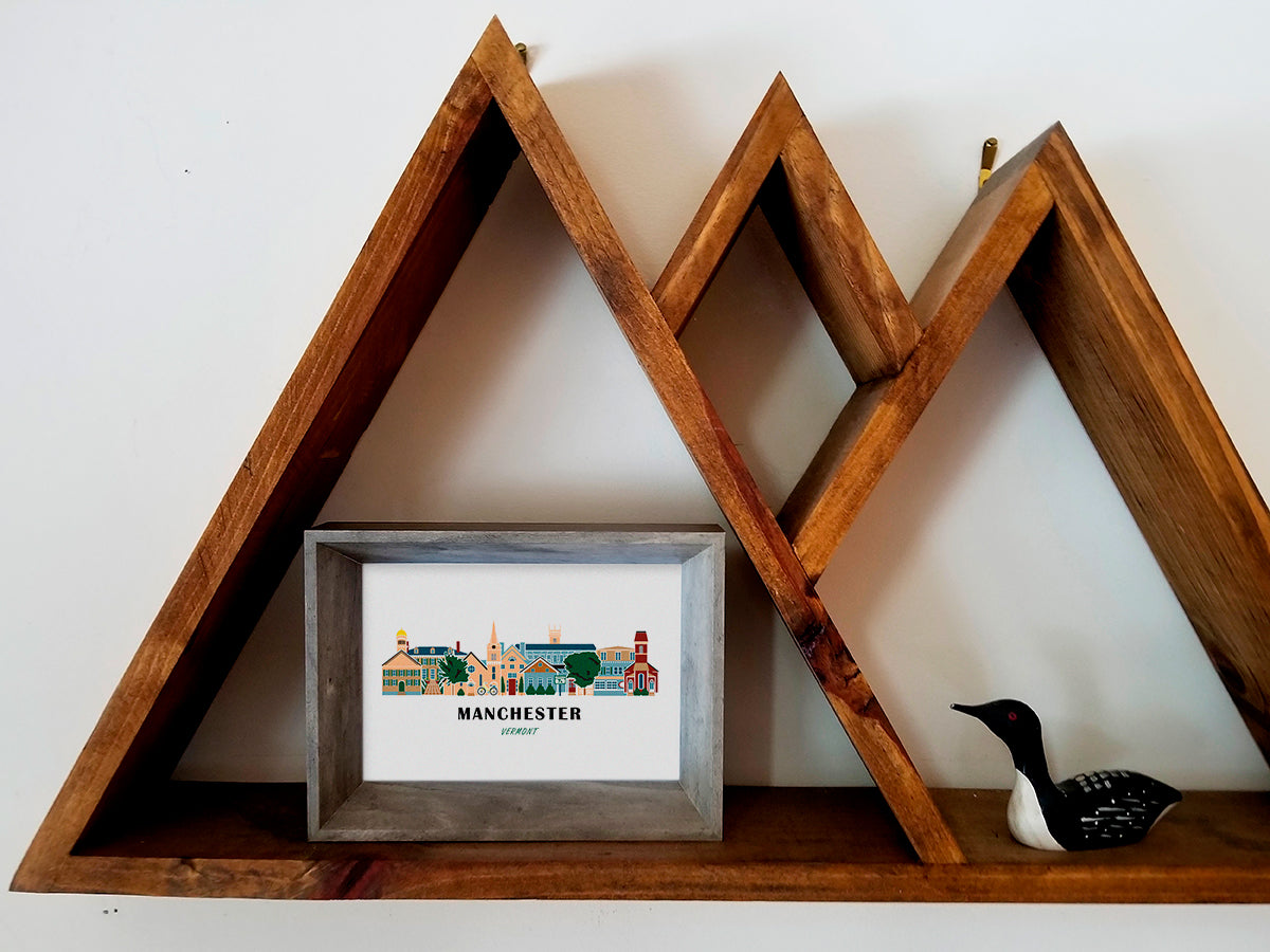 Framed art print on a wooden shelf of buildings arranged in a skyline with the text Manchester Vermont