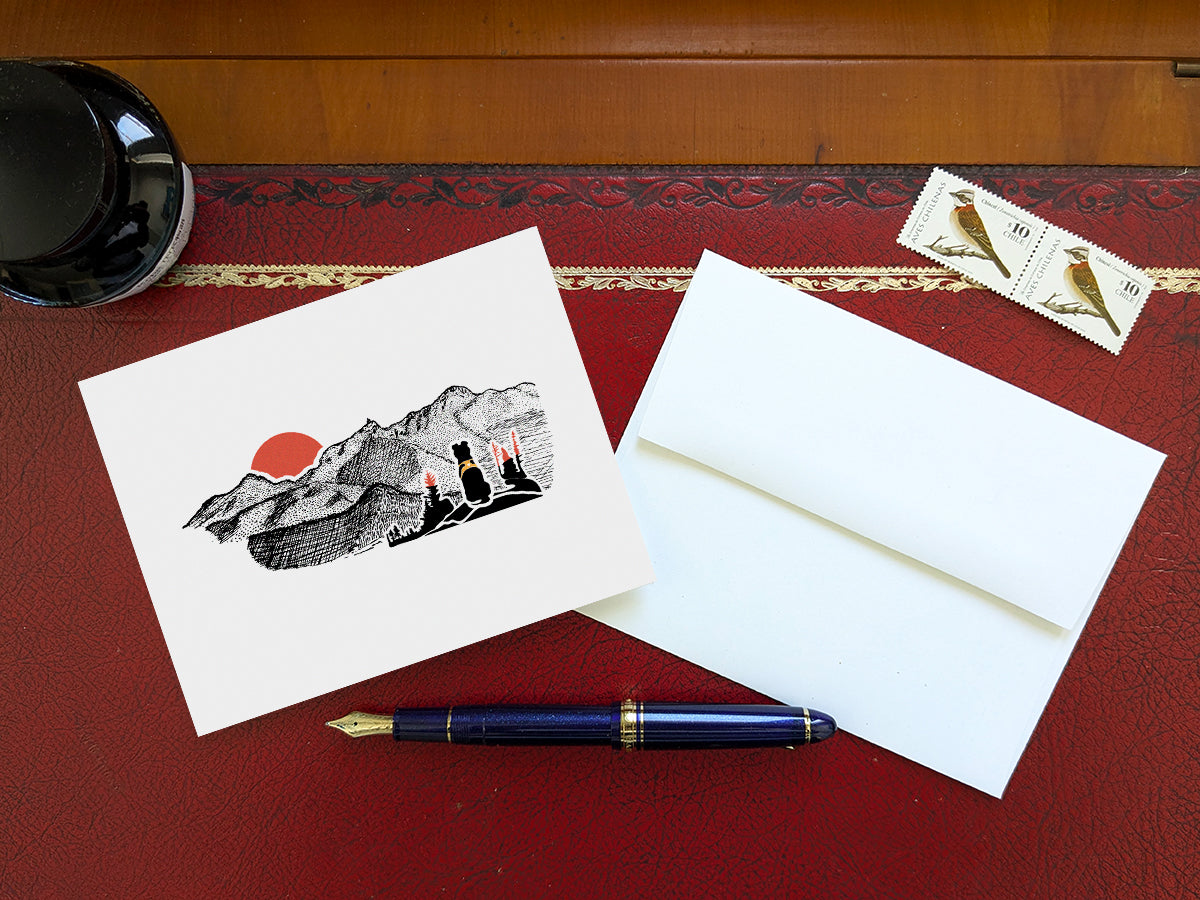 Greeting card with image of black dog looking at a red sunset behind mountains on a table with blue pen and envelope and stamps.