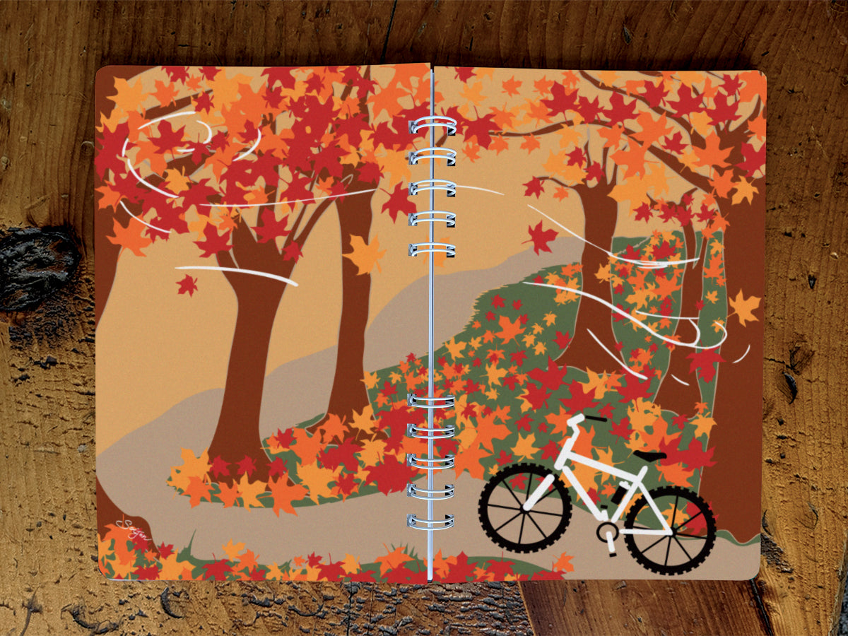 Notebook with autumn-themed cover featuring trees and a bicycle on a wooden surface