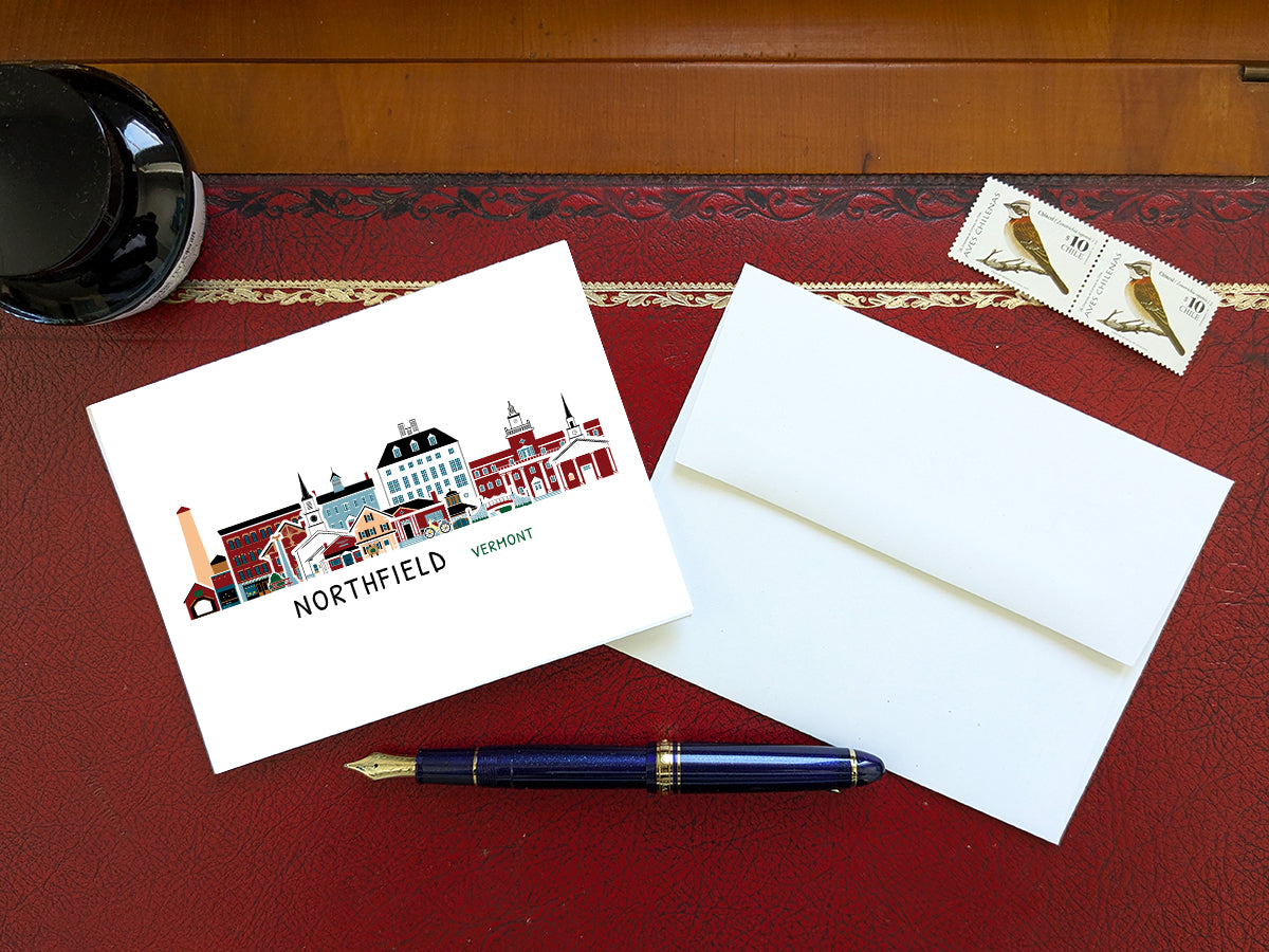 Greeting card with image of buildings arranged in a city skyline with the text Northfield Vermont on a red table with blue pen, stamps and envelope.