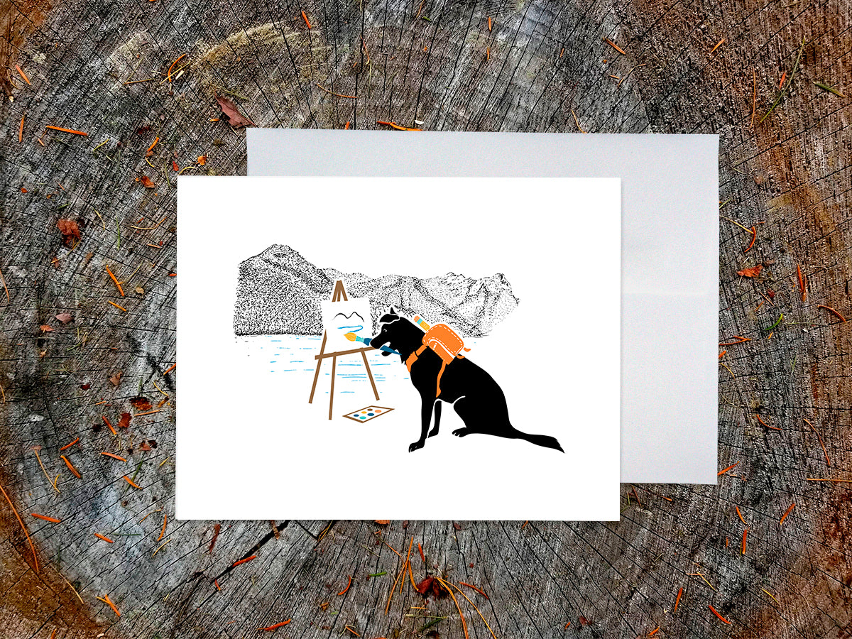 Greeting card with illustration of black dog painting in the mountains, with a white envelope placed on a wooden surface.