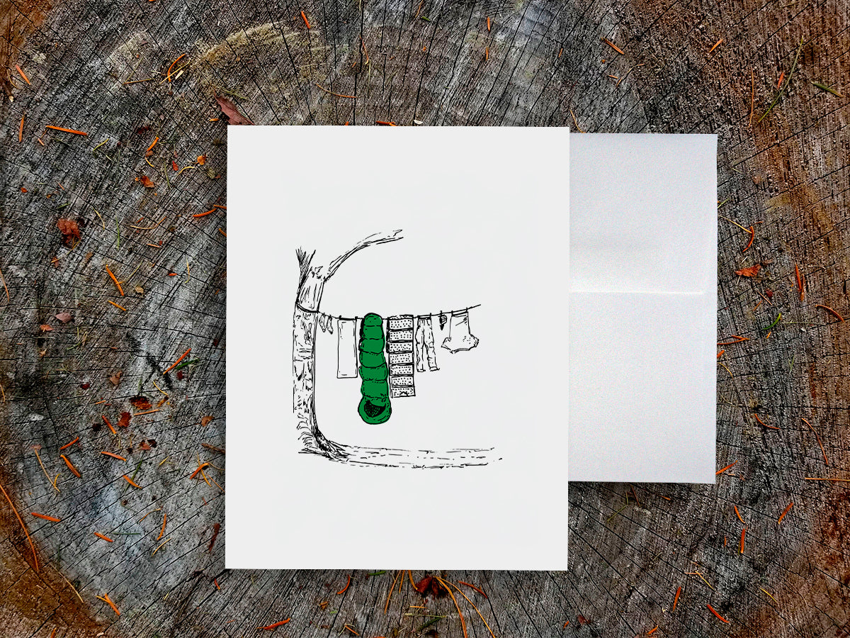 White card with a black ink drawing of laundry hanging from a tree on a wooden surface.