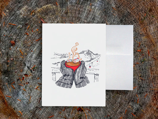 A greeting card with an illustration of a red, steaming cup of hot chocolate being held by big mittens, with mountains in the background, on a wood surface next to a white envelope.