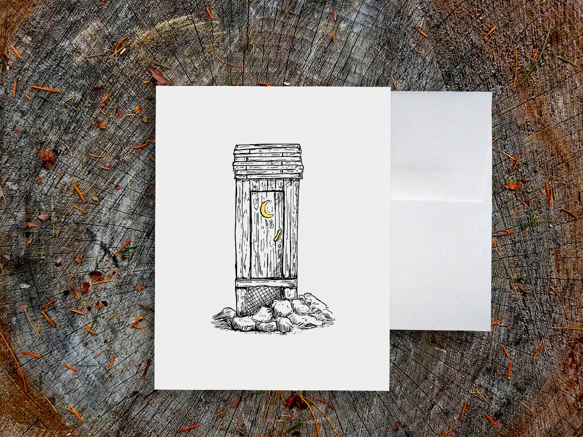 White card with a black ink drawing of an outhouse on a wooden surface.